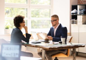 woman at desk talking to man holding clipboard in office setting