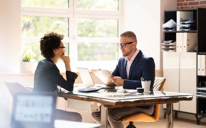 woman at desk talking to man holding clipboard in office setting